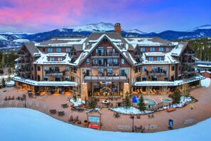 A large, multi-story hotel with a central courtyard, outdoor seating, and a view of snow-covered mountains. - Crystal Peak Condo | Spectacular Views, Steps To Peak 7, Hot Tubs, and Gondola on-site (Breckenridge)