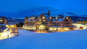 Un gran edificio de varios pisos con techo nevado y varios balcones, rodeado de un paisaje nevado y montañas al fondo.