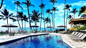 A poolside area with lounge chairs and umbrellas, palm trees, and a view of the ocean.