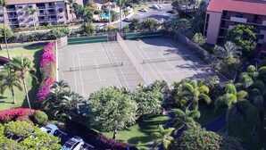 An aerial view of a tennis court surrounded by lush greenery and palm trees, with buildings in the background. - Maui Vista #2-101 Ground Floor, Pickle Ball Courts, Near Kamaole Beach Park (Kihei)