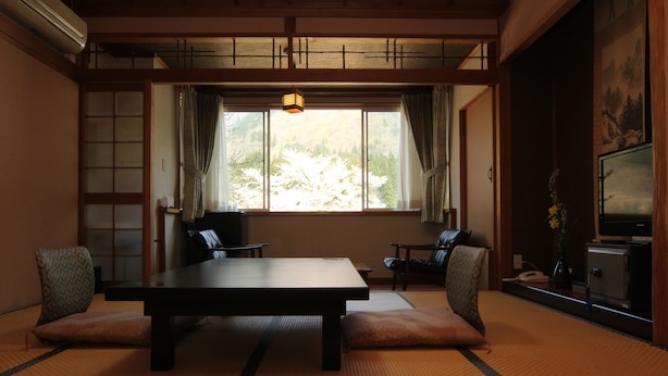 A traditional Japanese room with tatami flooring, a low table, cushions, a television, and a view of trees through a window. - Akao-Kan (Nanto)