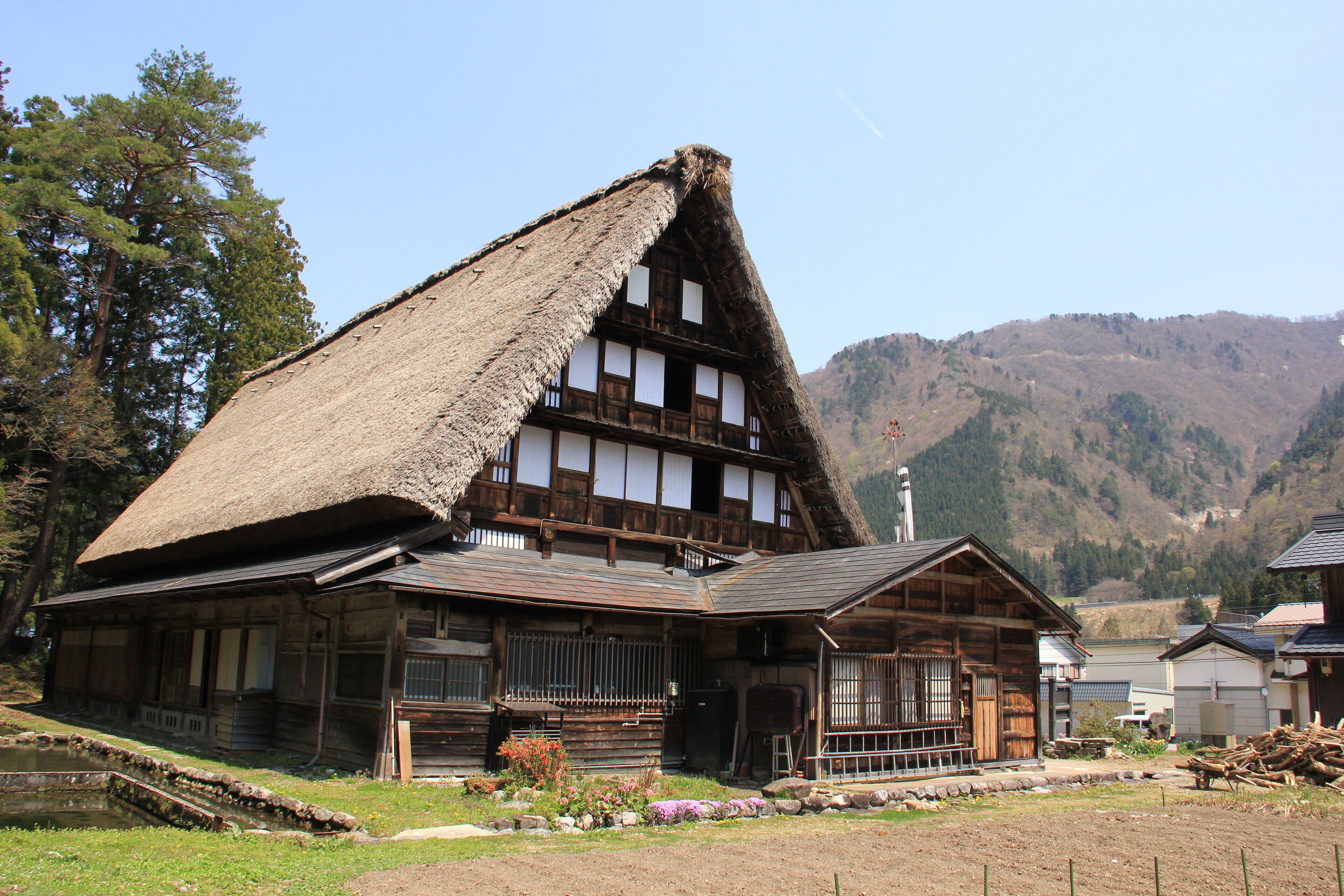 A traditional wooden house with a thatched roof, a wooden porch, and a small garden area with flowers and a pond.