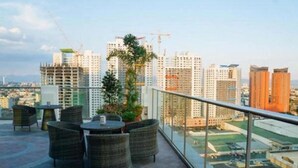 Eine Dachterrasse mit Korbmöbeln und Glasgeländer mit Blick auf eine Skyline mit Hochhäusern und Baukränen.