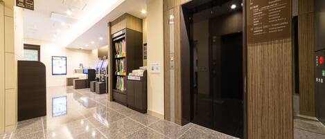 A modern hotel lobby with a reception desk, vending machines, and a digital information board.