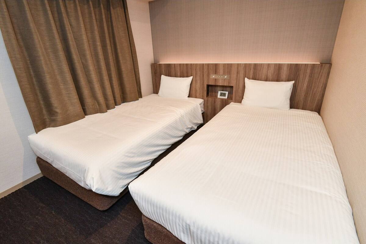 Une chambre d’hôtel avec deux lits simples, une tête de lit en bois, une petite table de chevet et un rideau.