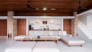 A modern living room with a sectional sofa, a coffee table, and a fireplace. There is a kitchen area with a stove and cabinets, and a staircase with a stone accent wall. - Villa Bliss (Kuta)
