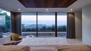 A modern bedroom with a large bed, a yellow chair, and a view of the countryside through large windows. - Villa Bliss (Kuta)
