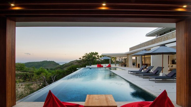 A modern outdoor pool area with a wooden table and red cushions, surrounded by lounge chairs and umbrellas, overlooking a hilly landscape. - Villa Bliss (Kuta)