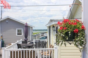 A deck with a table and chairs, a hanging flower basket, and a view of a body of water and vehicles in the background. - Premier, modern, fully renovated, Beach home...stroll to the beach in 3 minutes! (Hampton)