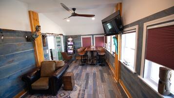 A cozy room with a wooden floor, a leather sofa, a dining table with chairs, a ceiling fan, and a television mounted on the wall.