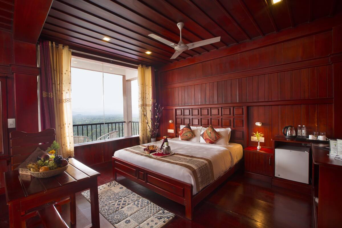 A hotel room with a wooden ceiling, a bed with a white comforter, a small desk with a bowl of fruit, and a balcony with a view.
