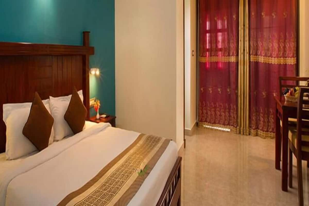 A hotel room with a wooden headboard, a bed with white and brown bedding, a nightstand with a lamp, and a view of a red curtain and a dining area.