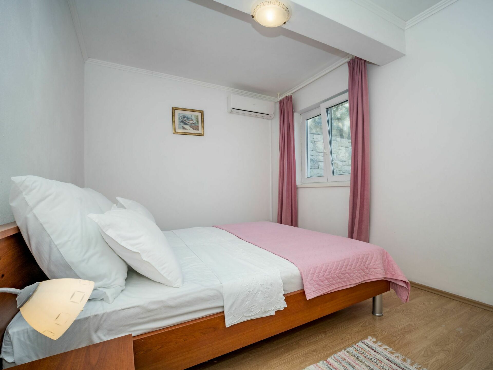 Een enkel bed met houten frame, witte beddengoed en een roze deken. Aan de muur hangt een klein, ingelijst beeld, een raam met roze gordijnen en een airconditioning.
