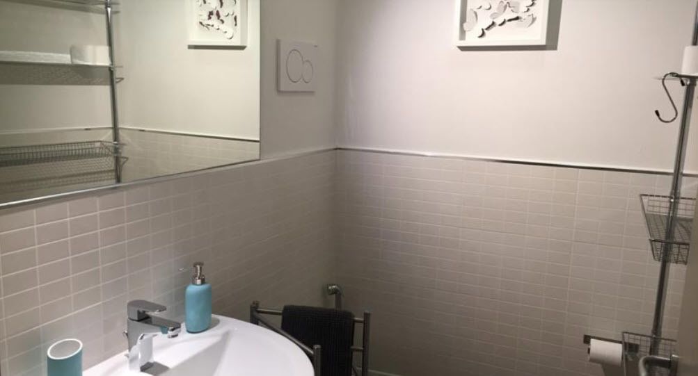 A bathroom with white tiled walls, a sink with a towel rack, a mirror, and a toilet paper holder.