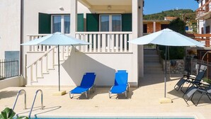 Ein Poolbereich mit zwei blauen Liegestühlen und Sonnenschirmen, eine Treppe, die zu einem Balkon führt, und ein freier Blick auf eine Hügelkulisse im Hintergrund.