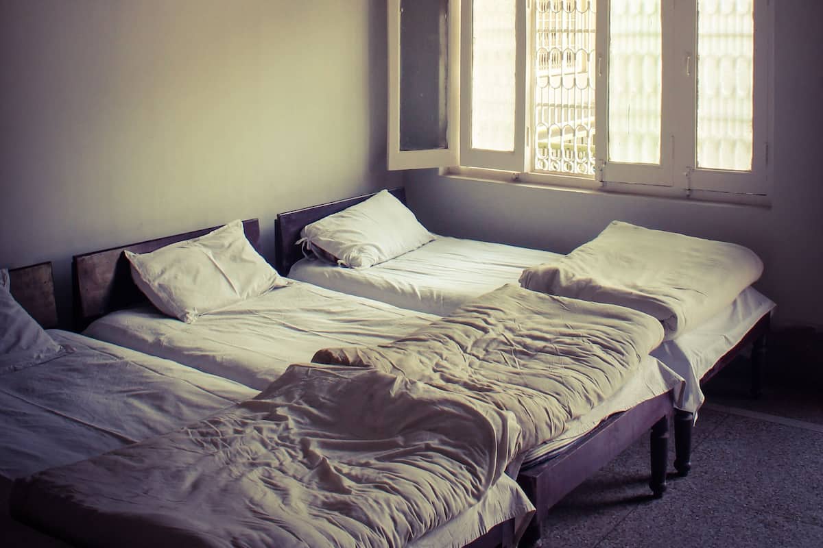 Quarto com duas camas, ventoinha no teto e janela com grades.