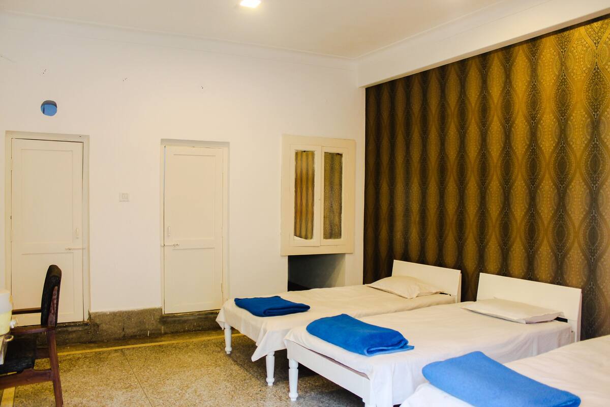 Quarto de hotel com três camas de solteiro, uma cadeira e uma janela grande com cortinas estampadas.