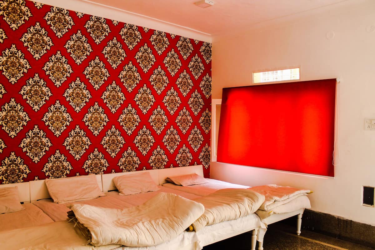 Quarto de hotel com papel de parede estampado, uma janela grande com cortina vermelha e uma cama com várias almofadas.
