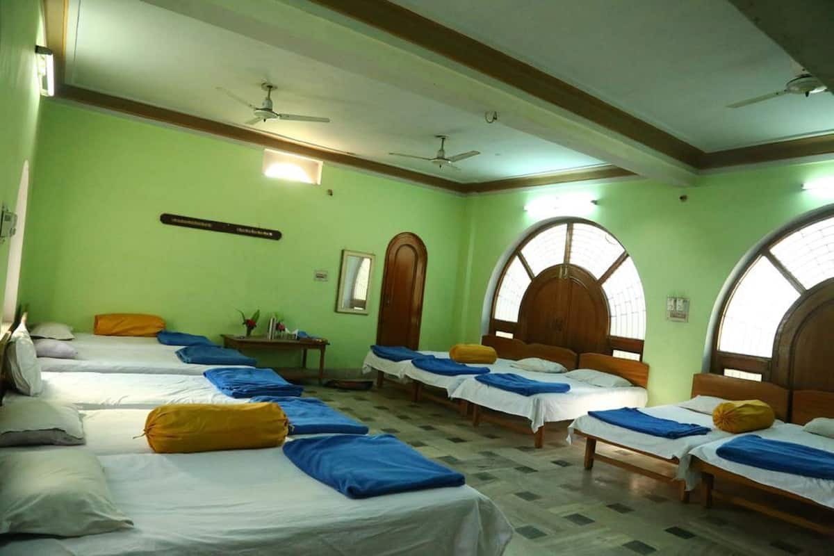 Quarto de hotel com várias camas, um ventilador de teto, uma pequena mesa com abajur e janelas em arco.
