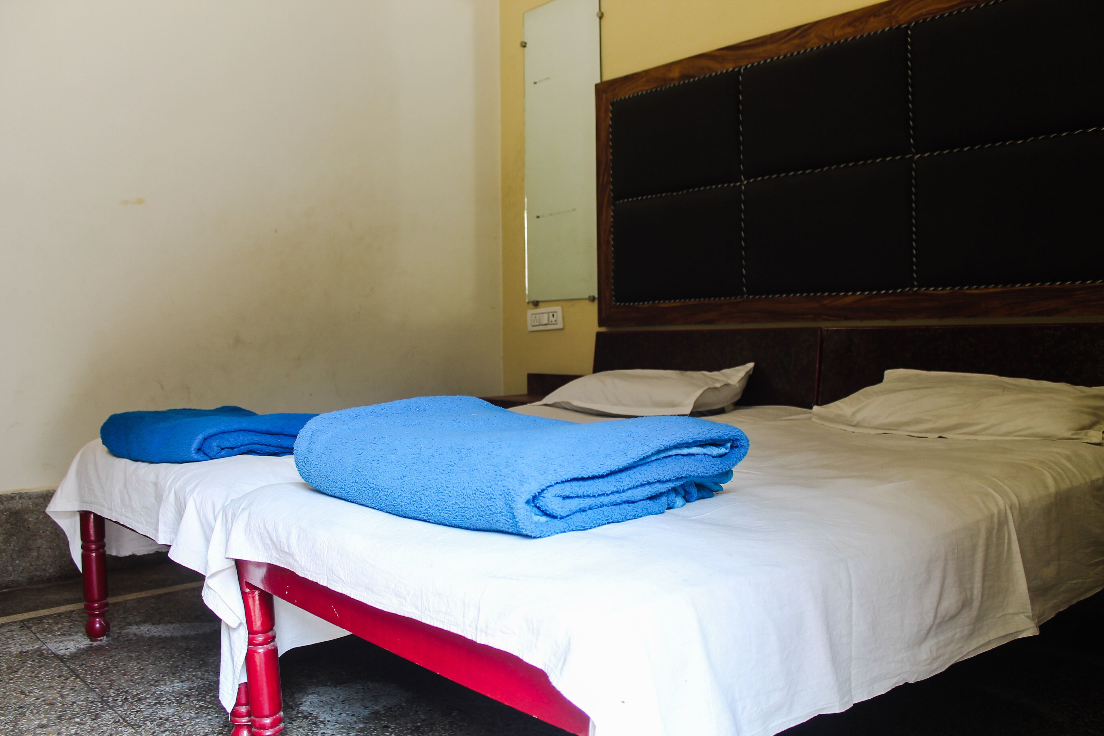 A single bed with a white sheet and two blue towels, a wooden headboard, and a plain wall.