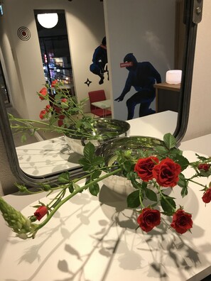 A vase with red roses on a white surface, a mirror reflecting a room with a red chair and a wall with a black silhouette. - bnb+ Ninja Otsuka - Hostel (Tokyo)