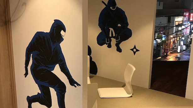 A modern interior with a minimalist design, featuring a wall with a large silhouette of a person in motion, a sleek white bench, and a view of a busy street through an open doorway. - bnb+ Ninja Otsuka - Hostel (Tokyo)