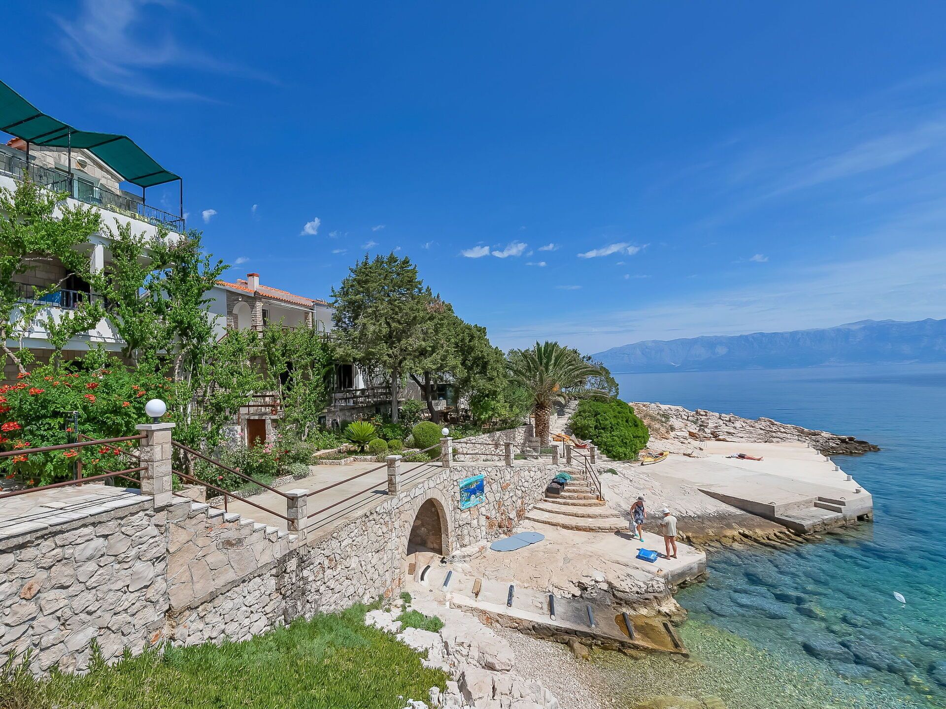 A coastal property with stone architecture, a clear view of the sea, and a stone staircase leading to a beach area.