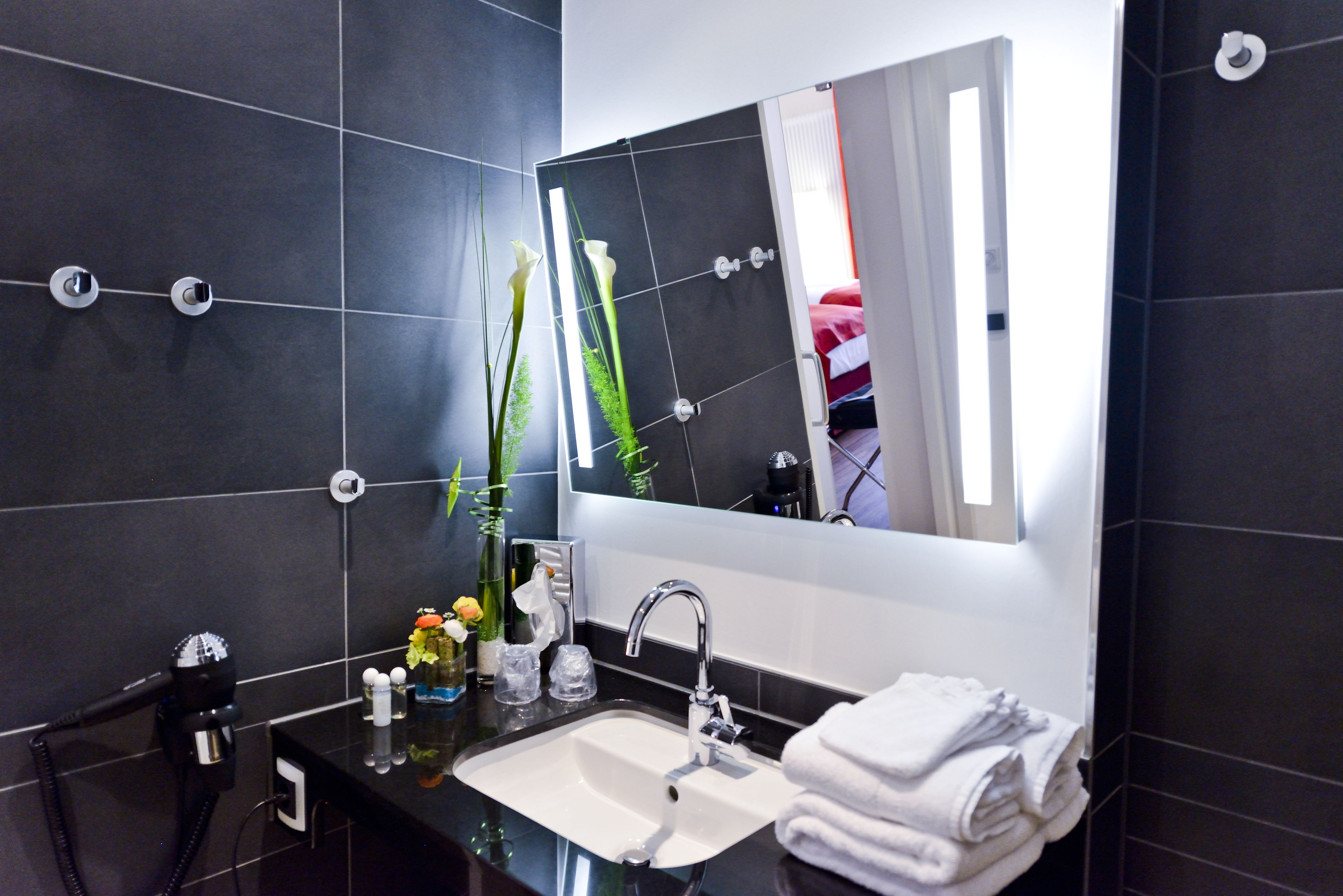 A modern bathroom with a black tiled wall, a large mirror, a white sink, and a towel rack.
