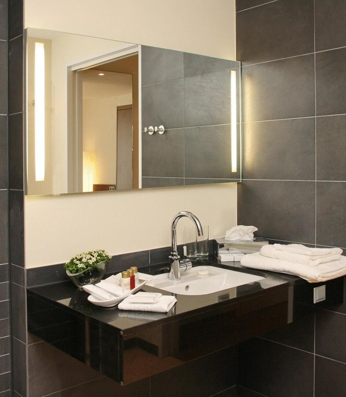 A modern bathroom with a dark tiled wall, a large mirror, a sink with a faucet, and a countertop with toiletries.