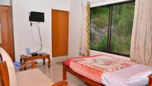 Un dormitorio con cama, una silla, una mesita, televisor montado en la pared y ventana con cortinas.