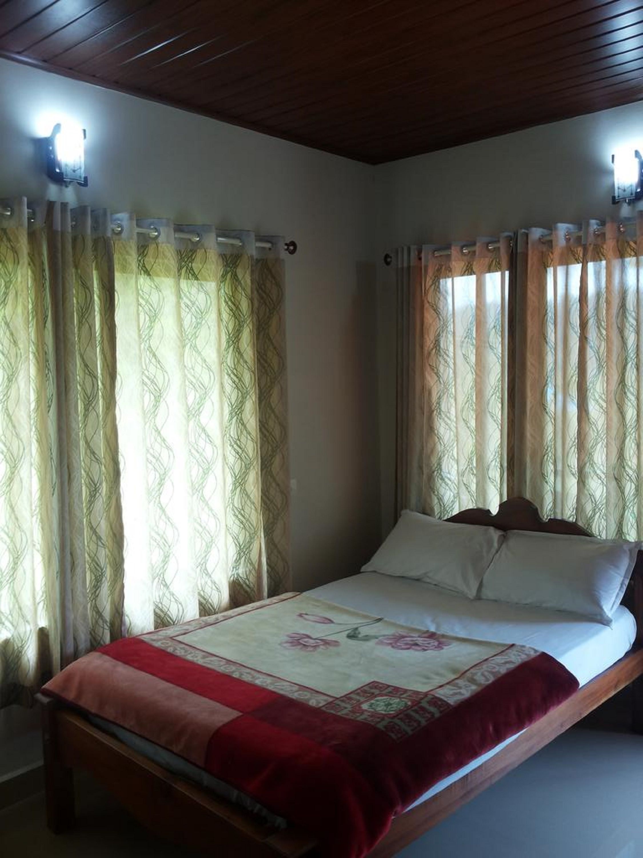 Un lit simple avec une couvre-lit rouge et blanche, deux oreillers blancs et une tête de lit en bois. La pièce est dotée de deux fenêtres avec des rideaux à motifs et le plafond est en bois.