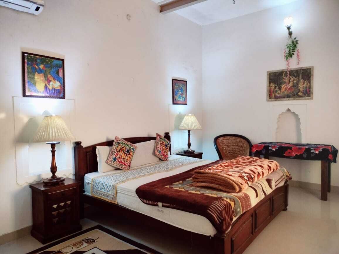 Une chambre avec un lit en bois, deux lampes de chevet, une table de chevet, une chaise, un bureau recouvert d’une nappe à motifs floraux et deux tableaux accrochés au mur.