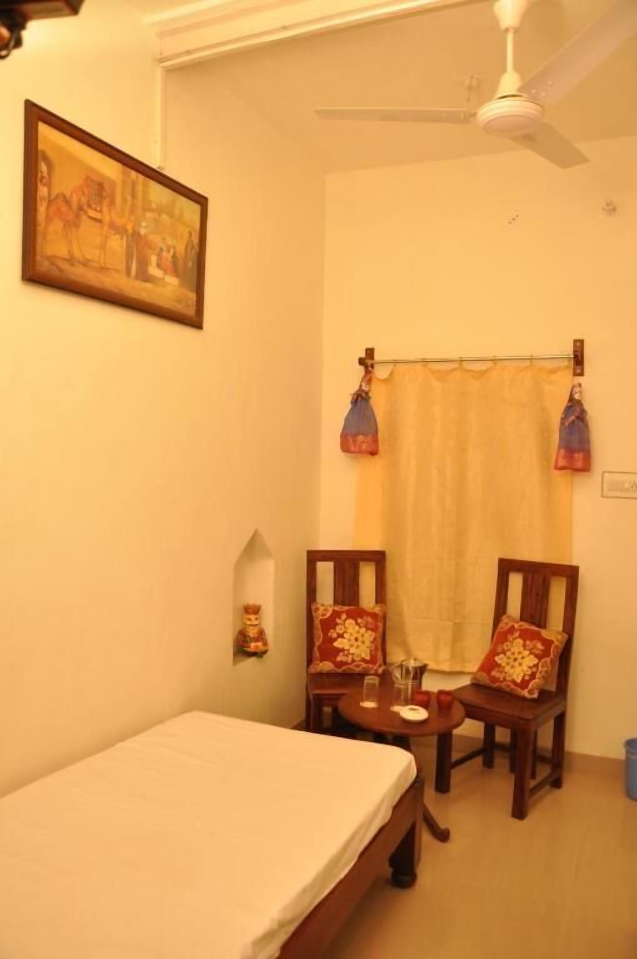 Una habitación pequeña con una cama, dos sillas, una mesita, un ventilador de techo y un cuadro en la pared.