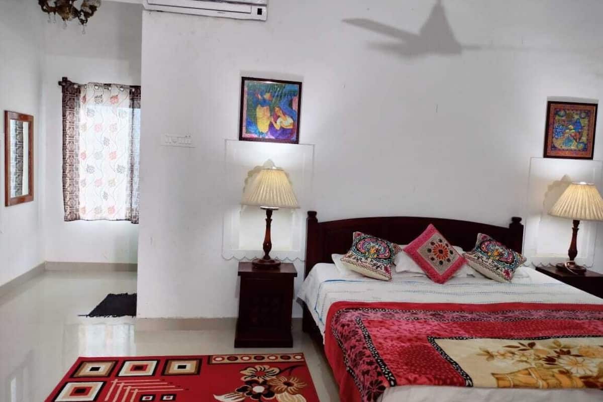 Un dormitorio con una cama, dos mesitas de noche con lámparas, una alfombra roja estampada y dos cuadros colgados en la pared.