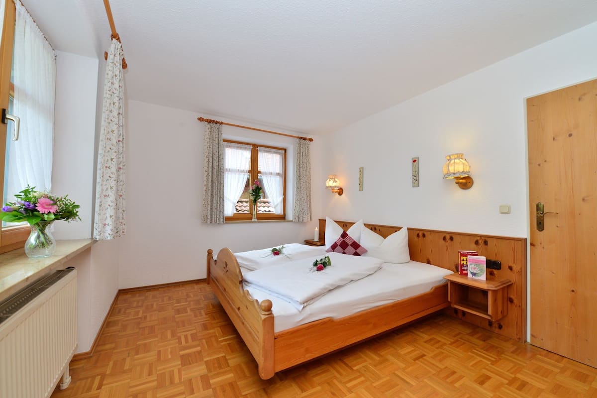 A bedroom with a wooden bed, a window with curtains, a bedside table with a book, and a wall-mounted light fixture.