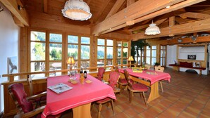A dining area with wooden tables and chairs, large windows with a view of the outdoors, and a high wooden ceiling. - Haus Jagdhof (Bad Hindelang)