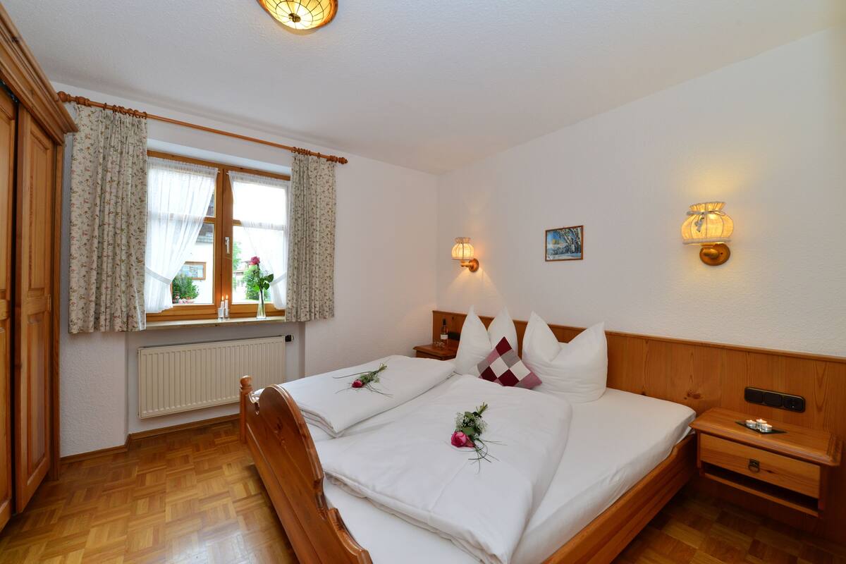 A hotel room with two beds, wooden headboards, a window with curtains, a radiator, and wall-mounted lamps.