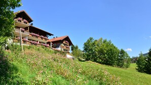A mountain lodge with balconies adorned with flowers, surrounded by lush greenery and a clear blue sky. - Haus Jagdhof (Bad Hindelang)