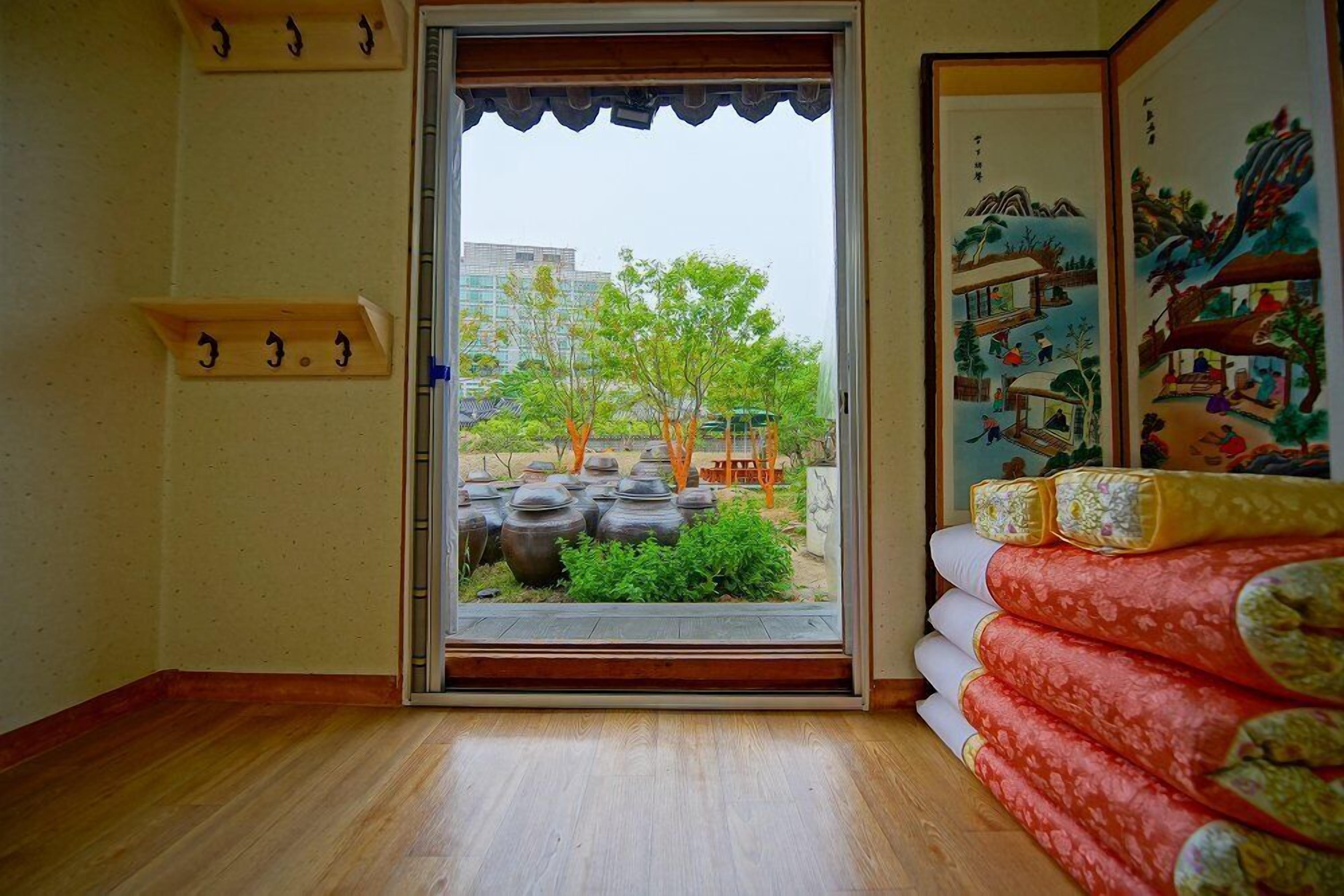 A room with a wooden floor, a sliding glass door leading to an outdoor area with potted plants and a view of buildings. There are wooden shelves with hooks on the wall and a stack of rolled-up mats on the floor.