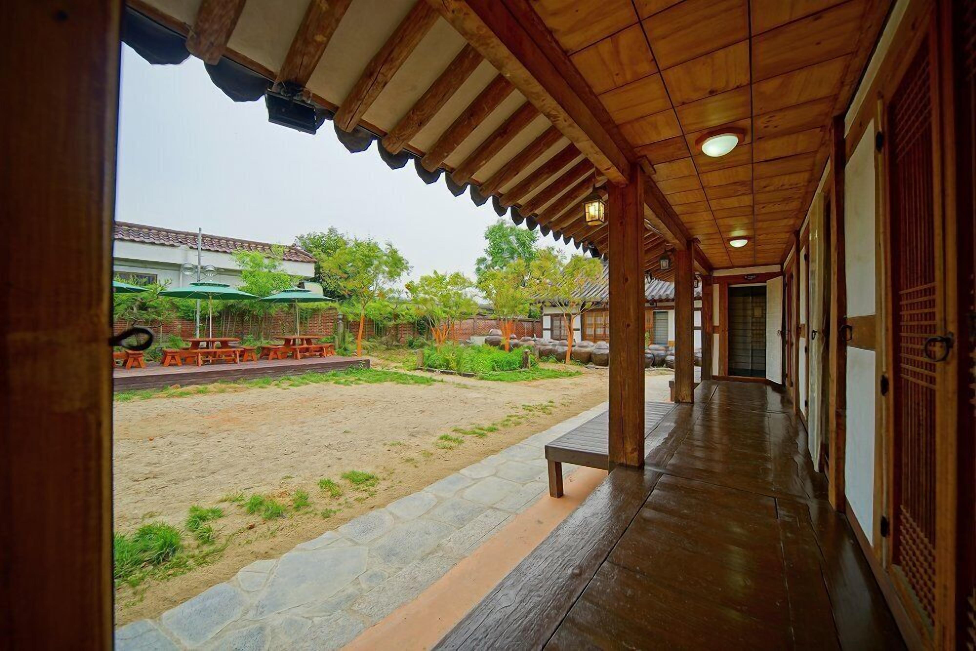 A covered walkway with a wooden ceiling and stone flooring, leading to a courtyard with outdoor seating and greenery.
