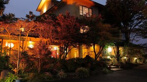 Uma casa japonesa tradicional com entrada bem iluminada, rodeada por um jardim com diversas plantas e pedras.