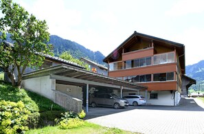 Un edificio de varios pisos con estacionamiento techado, montañas al fondo y un árbol en primer plano.