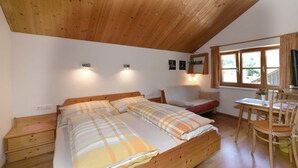 A bedroom with a wooden ceiling, a bed with plaid bedding, a bedside table, a chair, and a desk with a plant.