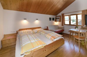 Une chambre avec un plafond en bois, un lit recouvert d’une couette à carreaux, une table de chevet, une chaise et un bureau agrémenté d’une plante.