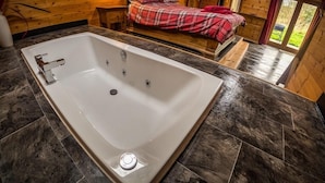 A modern bathroom with a large bathtub, wooden walls, and a bed with plaid bedding in the background. - New Forest Lodges (Wimborne)