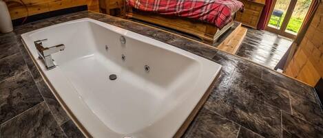 A modern bathroom with a large bathtub, wooden walls, and a bed with plaid bedding in the background.