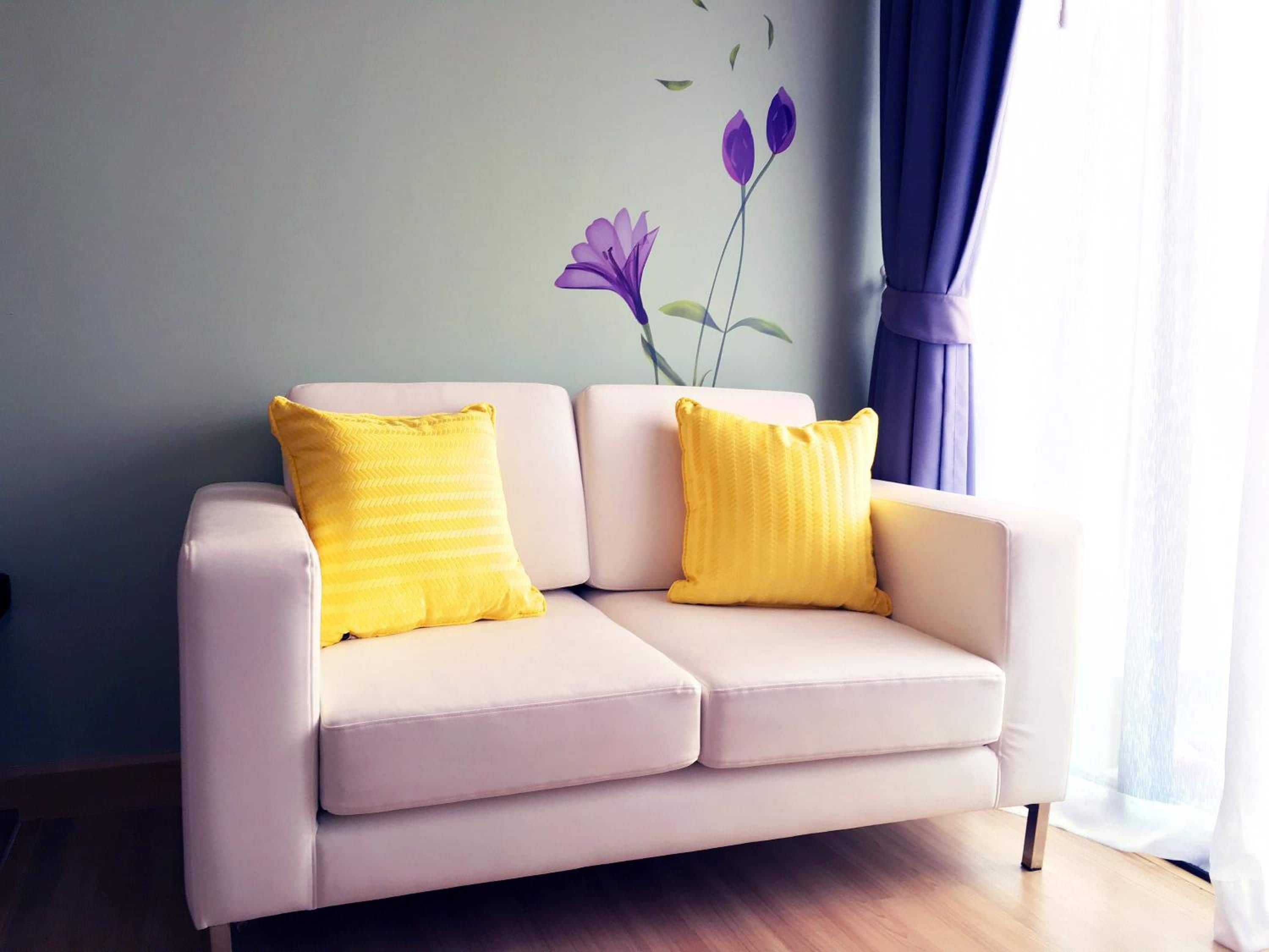Ein helles Sofa mit zwei gelben Kissen, platziert in einem Raum mit Holzboden und einer Wand mit floralem Wandbild.