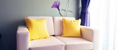 Un sofá de color claro con dos almohadones amarillos, ubicado en una habitación con piso de madera y un mural floral en la pared.