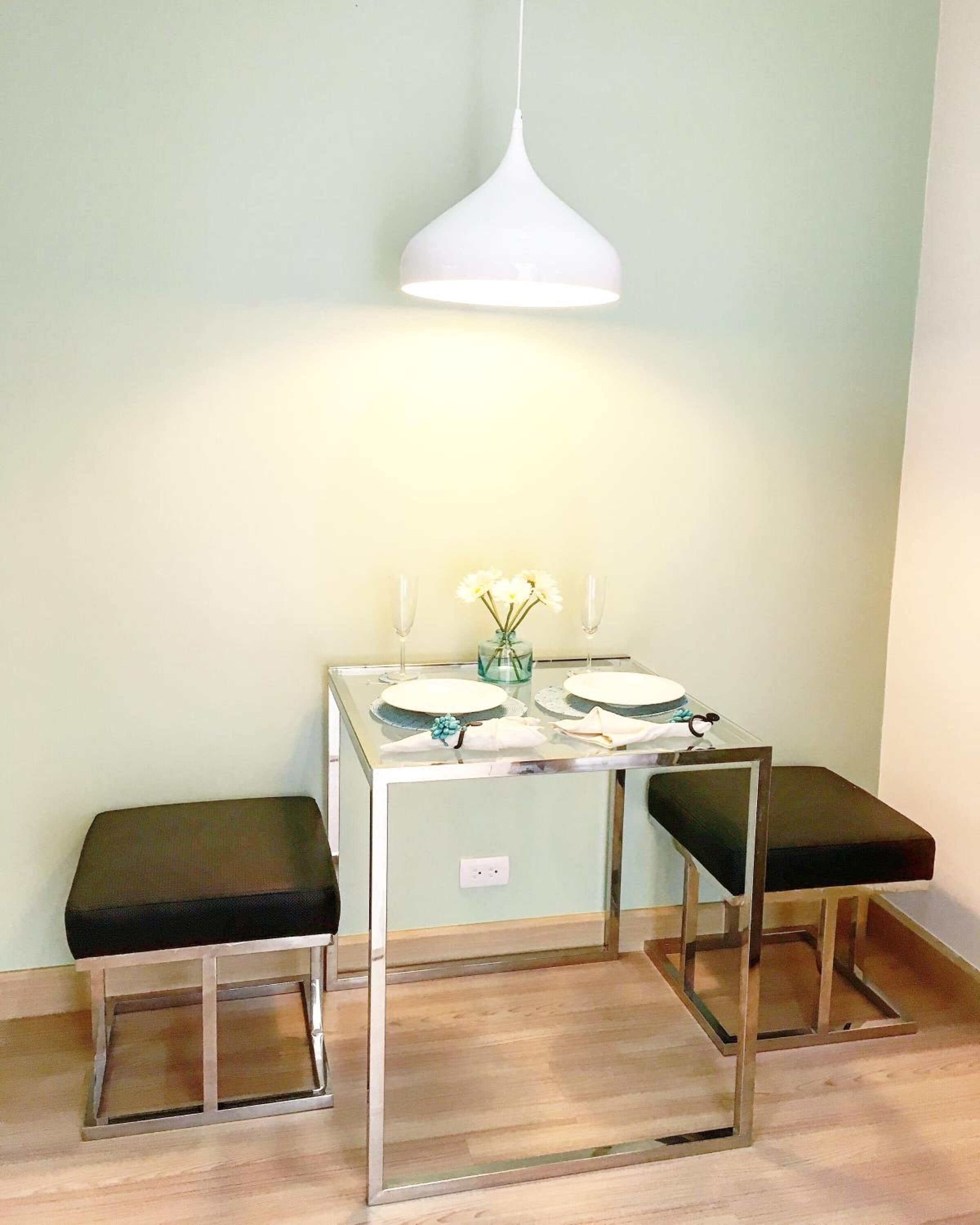 A small dining area with a table and two stools, a hanging pendant light, and a vase with flowers.