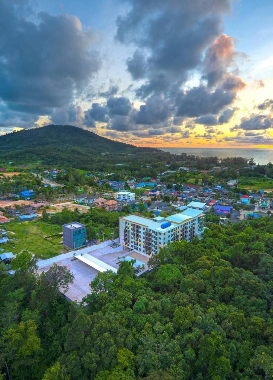A residential area with a mix of single and multi-story buildings, surrounded by dense greenery and a hilly backdrop.